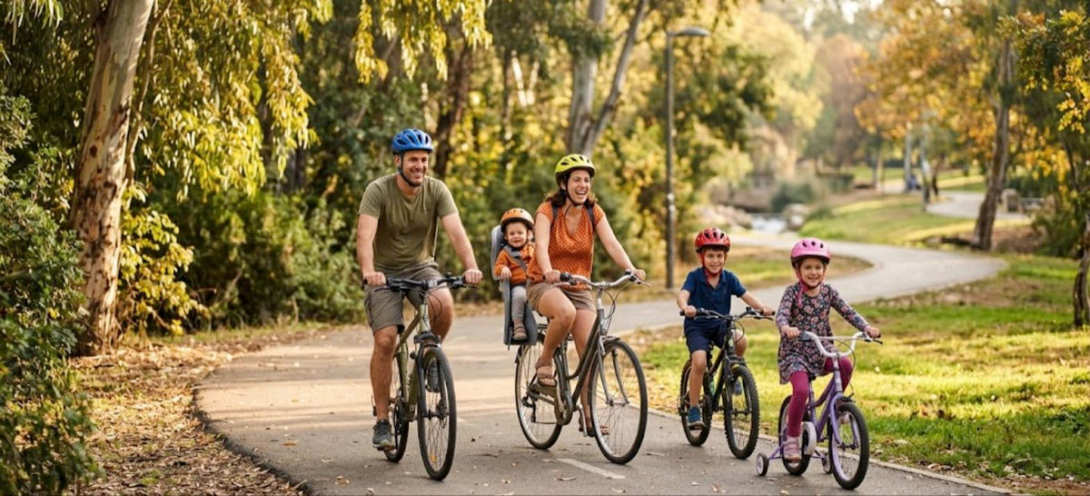 משפחה רוכבת יחד על אופניים בשביל פארק בבוקר סתווי בחול המועד סוכות, כולם חובשים קסדות, וילד קטן יושב בכיסא אחורי